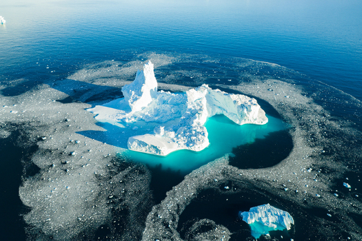 Aerial view of a large jagged iceberg surrounded by smaller ice fragments floating in the deep blue waters of the Arctic sea in Greenland. Aerial view of a large jagged iceberg surrounded by smaller ice fragments floating in the deep blue waters of the Arctic sea in Greenland.
