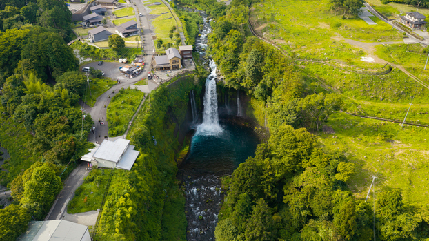 Aerial view of a waterfall cascading into a deep pool surrounded by dense green forest winding roads and small buildings in Japan. Aerial view of a waterfall cascading into a deep pool surrounded by dense green forest winding roads and small buildings in Japan.