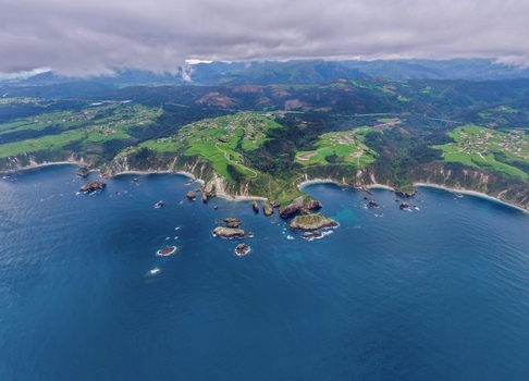 Aerial view of the rugged coastline of Cudillero featuring steep cliffs green meadows and rocky islets under a cloudy sky in Cudillero Asturias Spain. Aerial view of the rugged coastline of Cudillero featuring steep cliffs green meadows and rocky islets under a cloudy sky in Cudillero Asturias Spain.