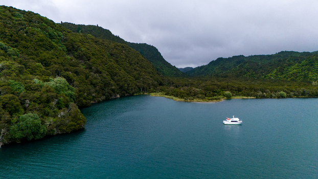 Aerial view of a serene lake reflecting the overcast sky with a single boat amidst the lush green forested hills Lake Tarawera New Zealand. Aerial view of a serene lake reflecting the overcast sky with a single boat amidst the lush green forested hills Lake Tarawera New Zealand.