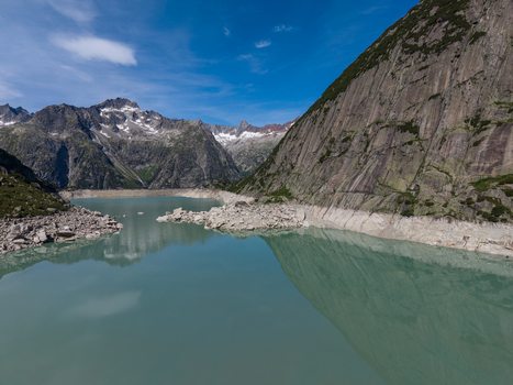 Aerial view of Gelmersee reservoir in the Swiss Alps showing low water levels rocky shoreline and surrounding mountain peaks under a clear blue sky in Switzerland.