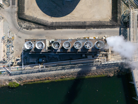Aerial view of an industrial cooling tower array with large fans and steam emissions situated near a waterway and infrastructure in Carson California United States.