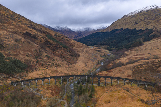 Aerial view of the Glenfinnan Viaduct curved railway bridge spanning a valley with snow capped mountains and a winding river Glenfinnan Scotland United Kingdom.