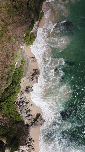 Aerial view of waves crashing against a rocky coastline with sandy beach and lush green cliffs in Indonesia.