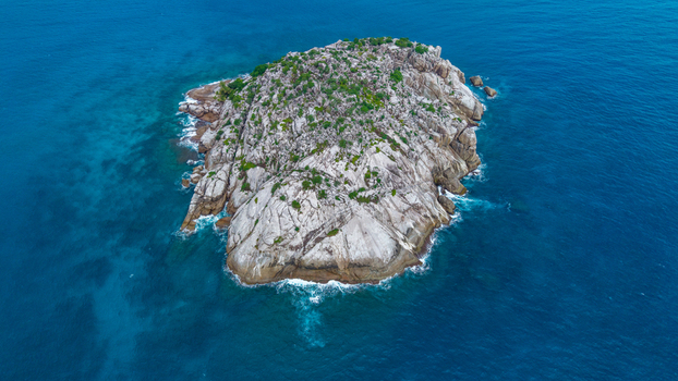 Aerial view of a small rocky island surrounded by deep blue ocean waters with crashing waves and sparse green vegetation on the granite surface in Grand Anse Village Grand Anse Mahe Seychelles. Aerial view of a small rocky island surrounded by deep blue ocean waters with crashing waves and sparse green vegetation on the granite surface in Grand Anse Village Grand Anse Mahe Seychelles.