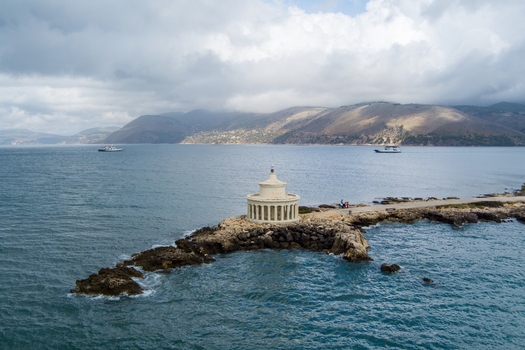Aerial view of the Saint Theodore Lantern lighthouse situated on a rocky peninsula surrounded by the blue sea with distant mountains under a cloudy sky in Argostoli Greece.