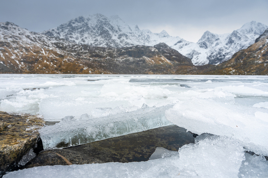 View of ice chunks on a frozen lake shore with snow capped mountains under an overcast sky in Leknes Lofoten Norway.