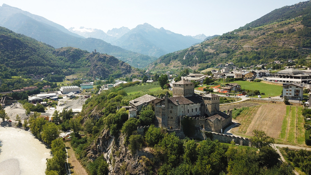 Aerial view of the Sarre Royal Castle on a rocky hill surrounded by lush greenery and a flowing river with the snow capped Alps in the background in Aosta Valley Aosta Valley Italy. Aerial view of the Sarre Royal Castle on a rocky hill surrounded by lush greenery and a flowing river with the snow capped Alps in the background in Aosta Valley Aosta Valley Italy.