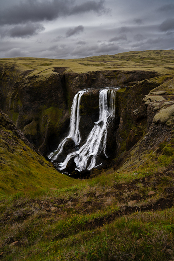 View of water cascades down a rugged cliff face contrasting with the green vegetation and dramatic sky creating a mesmerizing landscape Skaftárhreppur Iceland. View of water cascades down a rugged cliff face contrasting with the green vegetation and dramatic sky creating a mesmerizing landscape Skaftárhreppur Iceland.