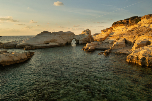 Aerial view of golden light bathing the rugged white and yellow rock formations and a natural arch contrasting with the deep turquoise sea under a serene sky Thira Thira Greece. Aerial view of golden light bathing the rugged white and yellow rock formations and a natural arch contrasting with the deep turquoise sea under a serene sky Thira Thira Greece.