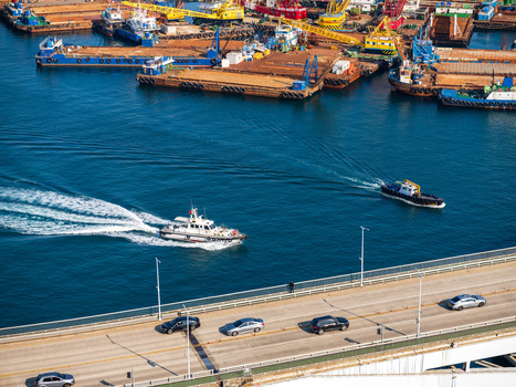 Aerial view of a bridge with cars a patrol boat navigating the blue water and industrial barges docked in the port area in Busan Busan South Korea. Aerial view of a bridge with cars a patrol boat navigating the blue water and industrial barges docked in the port area in Busan Busan South Korea.
