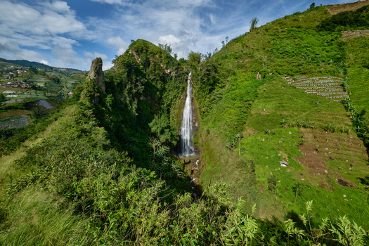 View of Curug Surodipo waterfall cascading down lush green cliffs surrounded by terraced agricultural fields under a blue sky in Temanggung Central Java Indonesia.