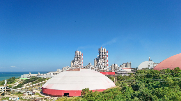 Aerial view of a large industrial cement plant featuring tall processing towers and dome shaped storage structures situated along the coast in Indonesia.