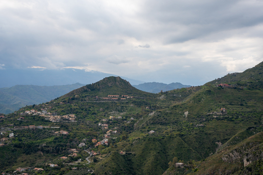 View of lush green hills dotted with terracotta roofed houses under a dramatic cloudy sky creating a serene yet striking landscape Taormina Sicily Italy. View of lush green hills dotted with terracotta roofed houses under a dramatic cloudy sky creating a serene yet striking landscape Taormina Sicily Italy.