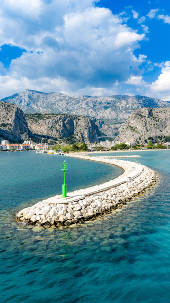 Aerial view of the curved stone breakwater and green beacon in the turquoise Adriatic Sea with the coastal town and rugged mountains in the background Omis Split Dalmatia County Croatia. Aerial view of the curved stone breakwater and green beacon in the turquoise Adriatic Sea with the coastal town and rugged mountains in the background Omis Split Dalmatia County Croatia.