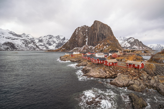 View of dramatic red cabins clinging to rocky shores against a backdrop of snow dusted mountains under a brooding sky Hamn&oslash;y Nordland Norway.