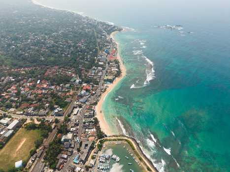 Aerial view of the turquoise coastline and sandy beach with fishing boats in the harbor and coastal buildings Hikkaduwa Southern Province Sri Lanka. Aerial view of the turquoise coastline and sandy beach with fishing boats in the harbor and coastal buildings Hikkaduwa Southern Province Sri Lanka.