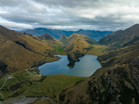 Aerial view of a serene lake nestled amidst the rugged golden mountains and green valleys under a cloudy sky Queenstown Otago Region New Zealand.