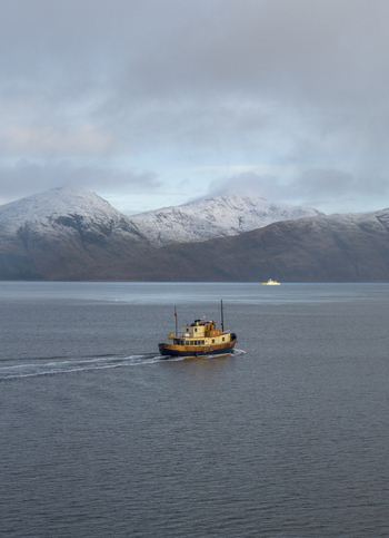 Aerial view of a vintage boat cruising on the calm waters of a loch with snow capped mountains in the background under a cloudy sky Scotland United Kingdom.
