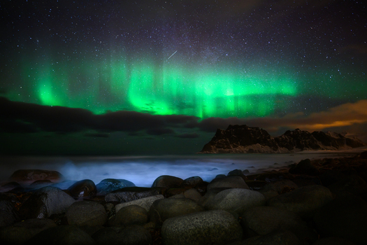 View of the vibrant green aurora borealis illuminating the night sky above a rocky beach with dark mountains in the background in Leknes Lofoten Norway.