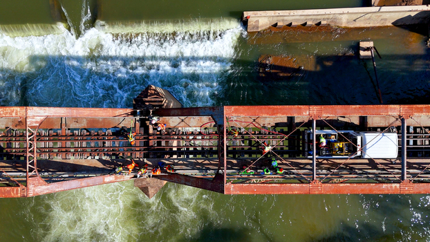 Aerial view of workers in high visibility gear performing maintenance on a rusty railway bridge over the Olifants River with water flowing over a weir in Hoedspruit Limpopo South Africa. Aerial view of workers in high visibility gear performing maintenance on a rusty railway bridge over the Olifants River with water flowing over a weir in Hoedspruit Limpopo South Africa.
