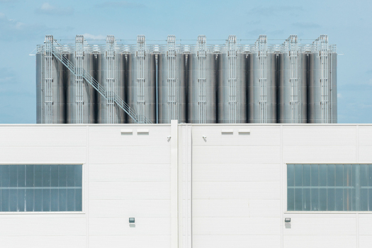 Aerial view of storage facilities for petrochemical products featuring rows of tall stainless steel tanks and a white industrial building under a clear blue sky in Poland.