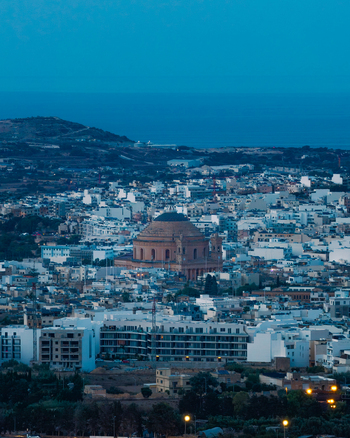 Aerial view of the Mosta Rotunda dome rising above the urban landscape of Mosta at dusk with white buildings and distant sea under a twilight sky. Mosta Northern Region Malta. Aerial view of the Mosta Rotunda dome rising above the urban landscape of Mosta at dusk with white buildings and distant sea under a twilight sky. Mosta Northern Region Malta.