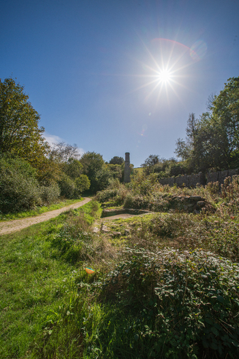 View of historic stone ruins and a chimney stack set within the lush landscape featuring a dirt path and bright sunlight in Tregargus Valley Cornwall United Kingdom. View of historic stone ruins and a chimney stack set within the lush landscape featuring a dirt path and bright sunlight in Tregargus Valley Cornwall United Kingdom.