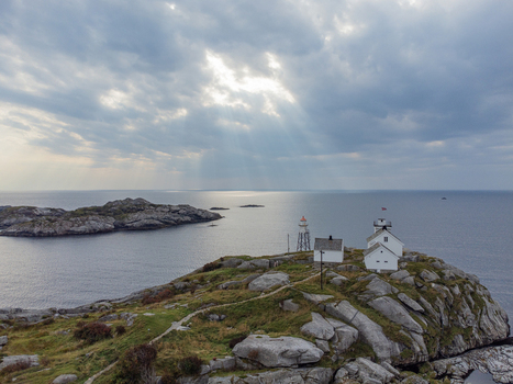 Aerial view of a small white lighthouse standing proudly on a rocky outcrop kissed by sunlight filtering through dramatic clouds Henningsvær Nordland Norway. Aerial view of a small white lighthouse standing proudly on a rocky outcrop kissed by sunlight filtering through dramatic clouds Henningsvær Nordland Norway.