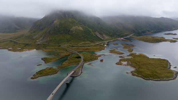 Aerial view of Lofoten bridge connecting islands surrounded by turquoise water lush green landscape and dramatic mountains under a cloudy sky in Nordland Norway.