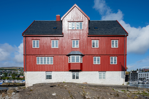 View of a traditional red wooden house with a white stone foundation situated on a rocky coastline under a clear blue sky Faroe Islands. View of a traditional red wooden house with a white stone foundation situated on a rocky coastline under a clear blue sky Faroe Islands.