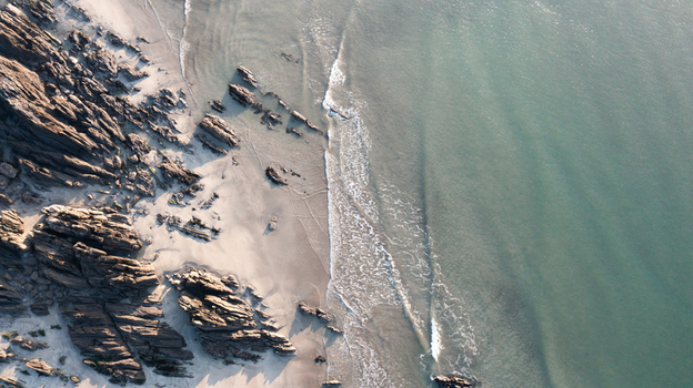 Aerial view of the rugged coastline with jagged rock formations and gentle waves washing over the sandy beach at Mothecombe Devon United Kingdom. Aerial view of the rugged coastline with jagged rock formations and gentle waves washing over the sandy beach at Mothecombe Devon United Kingdom.