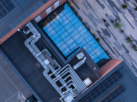 Aerial view of a modern building roof featuring complex HVAC ventilation ducts a large glass skylight and rows of solar panels with people walking on the adjacent paved plaza in Poland. Aerial view of a modern building roof featuring complex HVAC ventilation ducts a large glass skylight and rows of solar panels with people walking on the adjacent paved plaza in Poland.