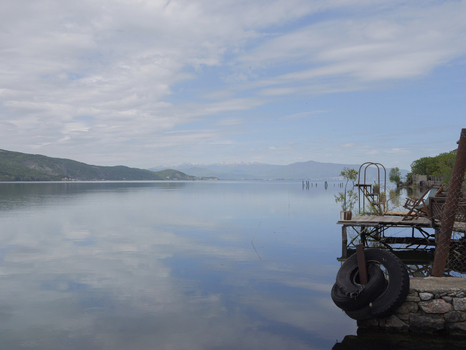 View of the calm waters of Lake Ohrid a small wooden pier a tire and distant mountains under a cloudy sky in Lin Korce County Albania.