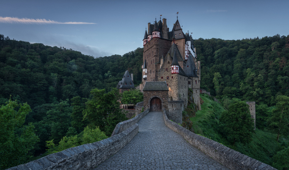 View of Eltz Castle a medieval fortress with stone towers and timber framed architecture situated on a rocky outcrop surrounded by a dense forest at dusk in Germany.
