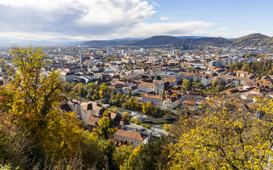 View of the cityscape unfolds with a tapestry of red tiled roofs golden trees and distant mountains under a bright sky Graz Styria Austria. View of the cityscape unfolds with a tapestry of red tiled roofs golden trees and distant mountains under a bright sky Graz Styria Austria.