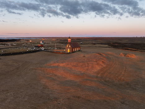 Aerial view of Hvalsneskirkja church standing in a vast golden lit landscape with a nearby cemetery under a soft sunset sky Sudurnesjabaer Iceland. Aerial view of Hvalsneskirkja church standing in a vast golden lit landscape with a nearby cemetery under a soft sunset sky Sudurnesjabaer Iceland.