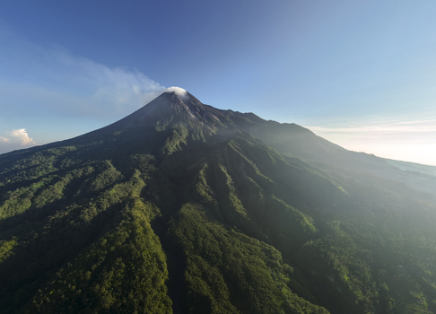 Aerial view of Mount Merapi volcano with steam rising from the crater above lush green forested slopes and Bunker Kaliadem in Kecamatan Pakem Daerah Istimewa Yogya Indonesia. Aerial view of Mount Merapi volcano with steam rising from the crater above lush green forested slopes and Bunker Kaliadem in Kecamatan Pakem Daerah Istimewa Yogya Indonesia.