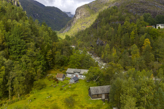 Aerial view of traditional wooden farmhouses nestled in a lush green valley with a flowing river and steep forested mountains in Fjaera Vestland Norway. Aerial view of traditional wooden farmhouses nestled in a lush green valley with a flowing river and steep forested mountains in Fjaera Vestland Norway.