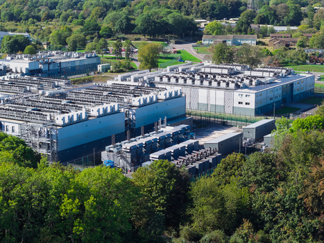 Aerial view of a large data center facility with industrial cooling units on the roof surrounded by lush green trees and vegetation in the United Kingdom.