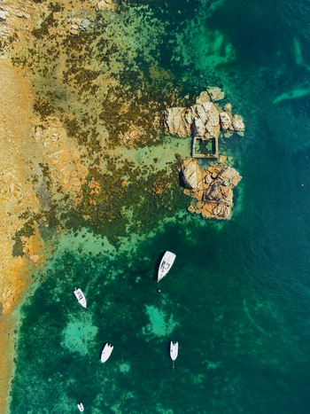 Aerial view of the rocky coast with white boats anchored in clear turquoise water featuring granite formations and seaweed covered shallows in Ile de Brehat Brittany France.