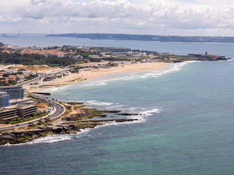 Aerial view of Carcavelos beach with its long sandy shore turquoise waves and the Sao Juliao da Barra Fort in the distance under a cloudy sky in Lisbon Portugal.