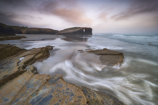 View of dramatic rock arches and cliffs at As Catedrais Beach with waves crashing on the sandy shore under a cloudy sunset sky Galicia Spain. View of dramatic rock arches and cliffs at As Catedrais Beach with waves crashing on the sandy shore under a cloudy sunset sky Galicia Spain.