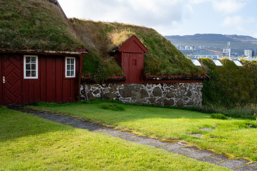 View of a traditional red wooden house with a grass roof and stone foundation set in a green landscape in the Faroe Islands. View of a traditional red wooden house with a grass roof and stone foundation set in a green landscape in the Faroe Islands.