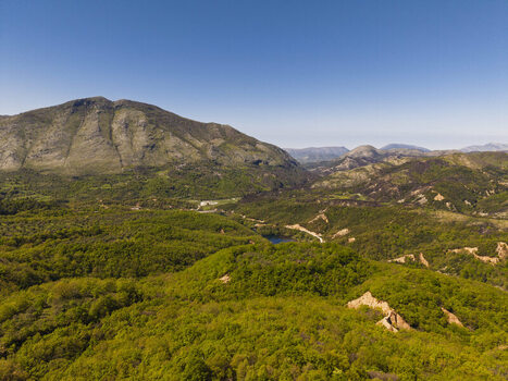 Aerial view of Muzine landscape featuring lush green forests rugged mountain slopes and a small lake nestled in the valley under a clear blue sky in Muzina Qarku i Vlores Albania. Aerial view of Muzine landscape featuring lush green forests rugged mountain slopes and a small lake nestled in the valley under a clear blue sky in Muzina Qarku i Vlores Albania.