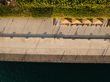 Aerial view of the waterfront promenade on the Island of Ortigia with lush green trees cafe umbrellas and long shadows cast on the stone pavement in Syracuse Sicily Italy. Aerial view of the waterfront promenade on the Island of Ortigia with lush green trees cafe umbrellas and long shadows cast on the stone pavement in Syracuse Sicily Italy.