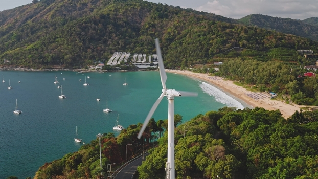 Aerial view of a wind turbine on a lush green hill overlooking the turquoise waters of Nai Harn Beach with sailboats anchored in the bay in Phuket Phuket Thailand. Aerial view of a wind turbine on a lush green hill overlooking the turquoise waters of Nai Harn Beach with sailboats anchored in the bay in Phuket Phuket Thailand.