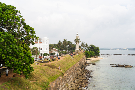 View of the historic Galle Fort Lighthouse and stone ramparts with people walking along the coastal fortifications under a cloudy sky Galle Southern Province Sri Lanka.