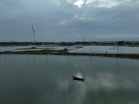 Aerial view of the Khurushkul area featuring a wind turbine calm water and a small boat with people under a cloudy sky in Cox s Bazar Chittagong Division Bangladesh. Aerial view of the Khurushkul area featuring a wind turbine calm water and a small boat with people under a cloudy sky in Cox s Bazar Chittagong Division Bangladesh.