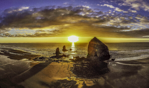 Aerial view of Haystack Rock at sunset with golden light reflecting on the wet sand and people walking along the shore Cannon Beach Oregon United States.